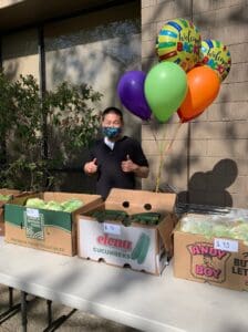 A Market Day volunteer gives thumbs up to the camera while standing behind a table full of produce and with balloons behind them