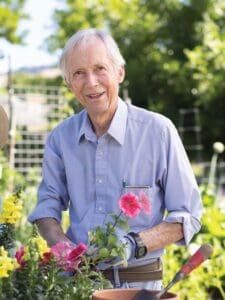 An older man stands in a garden with a pink flowering plant sitting on a table in front of him.