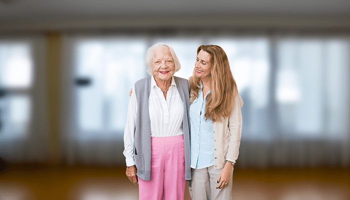 An older woman and a middle aged woman stand together in front of a blurred background.